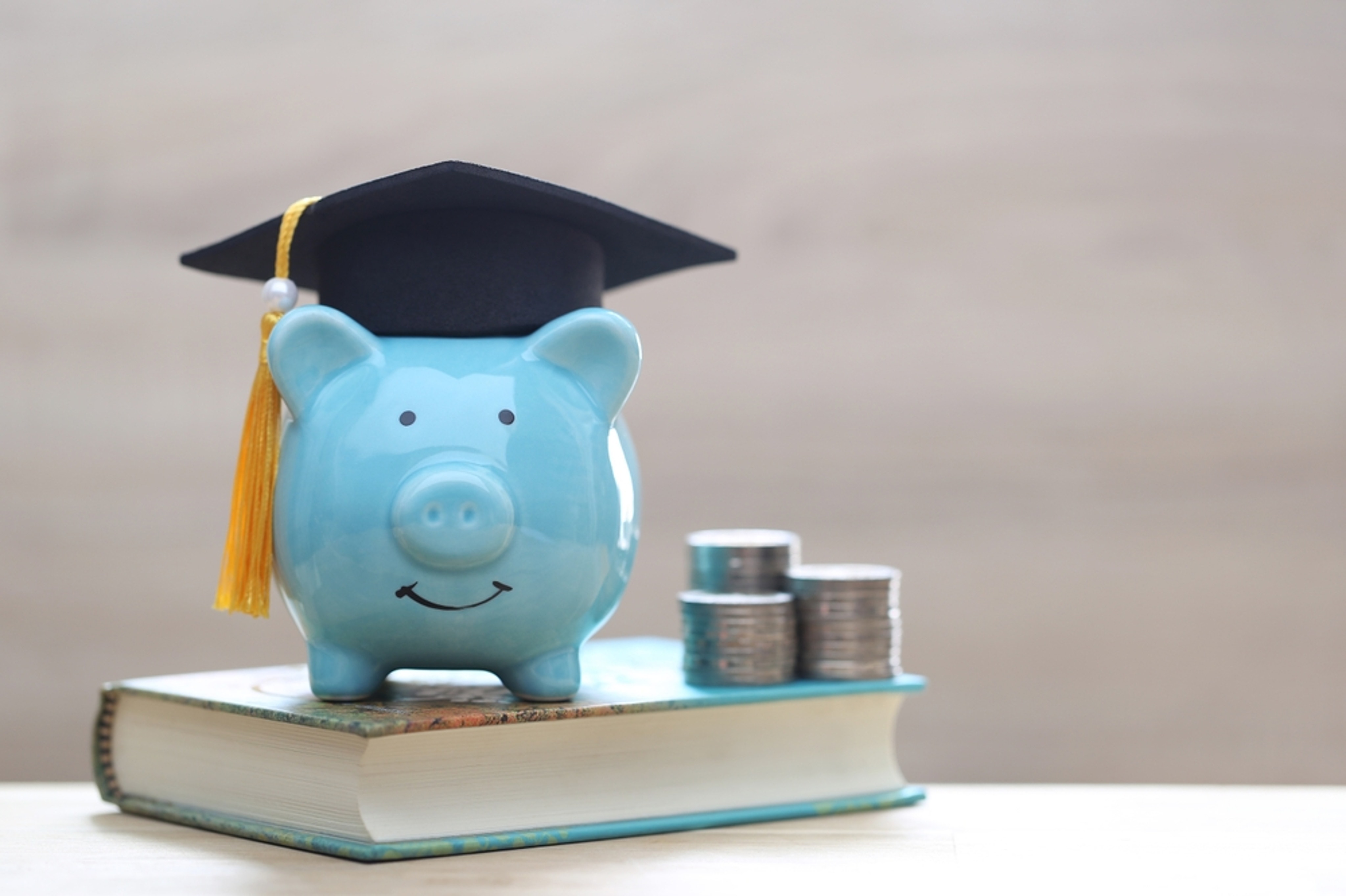 Graduation,Hat,On,Blue,Piggy,Bank,With,Stack,Of,Coins
